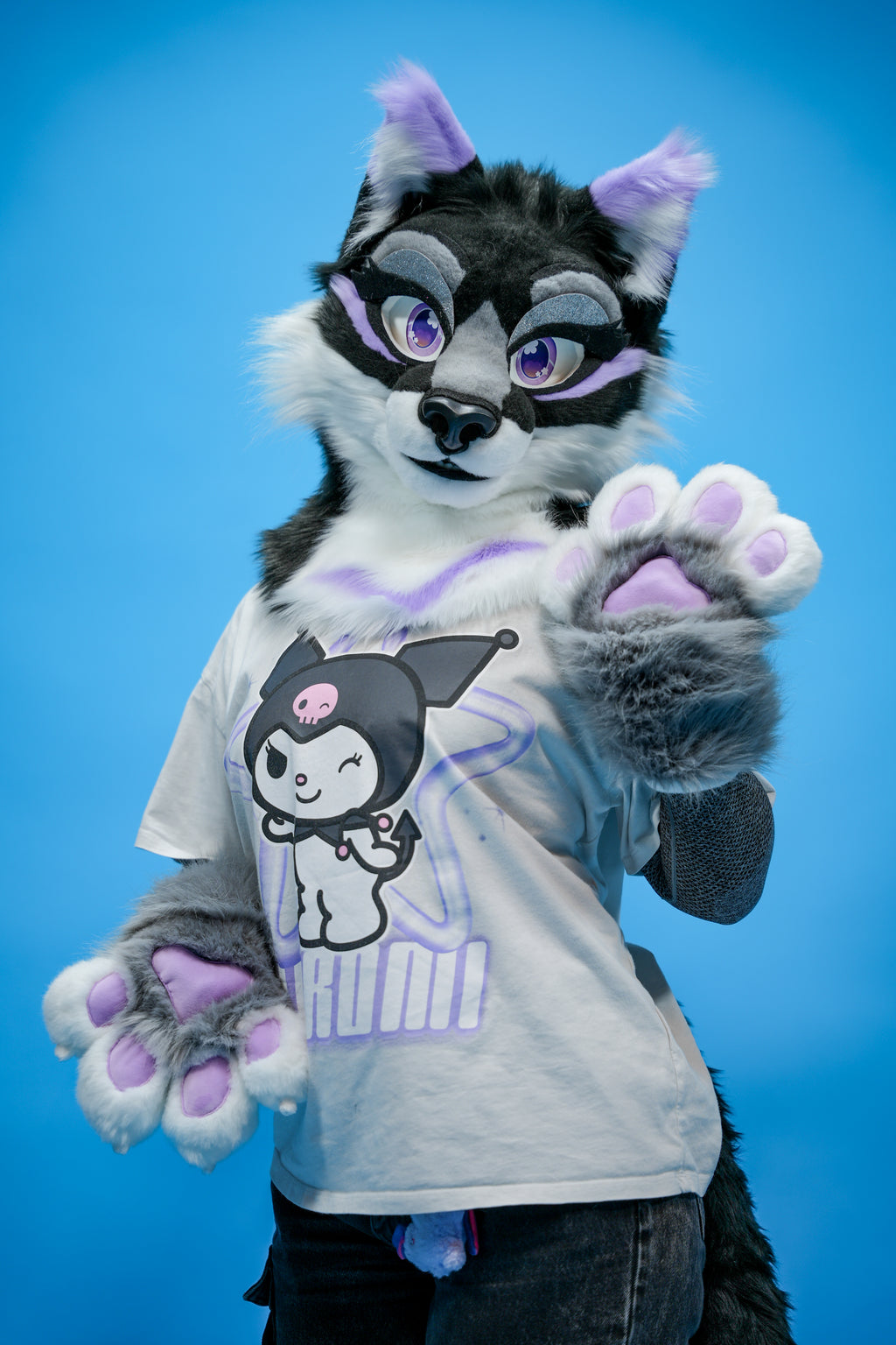 Person in a black and gray fursuit with purple accents wearing a white t-shirt with a graphic design on a blue background