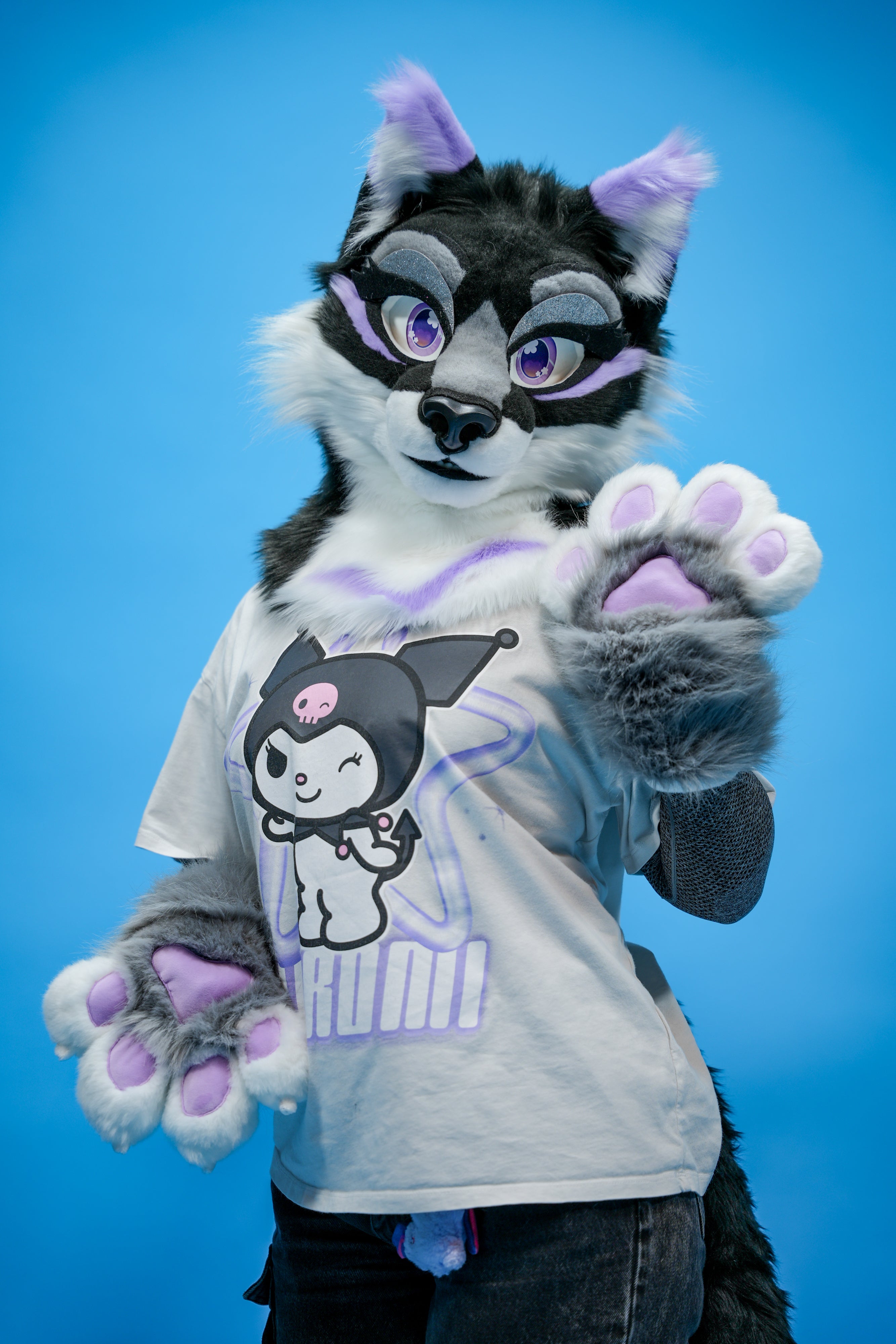 Person in a black and gray fursuit with purple accents wearing a white t-shirt with a graphic design on a blue background